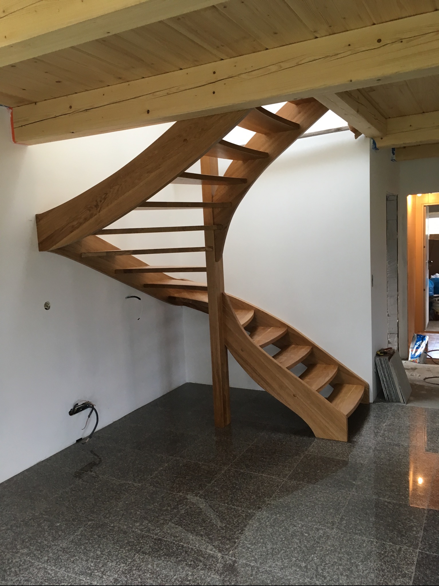 Kręte, jasne, drewniane schody bez poręczy, w trakcie montażu, z widocznymi przewodami elektrycznymi wystającymi ze ściany, na tle białej ściany i szarej, błyszczącej podłogi z granitu.