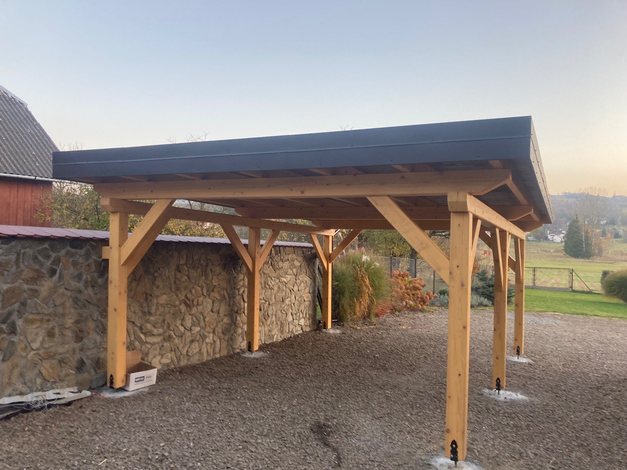 Drewniana pergola z płaskim, ciemnym dachem, widok na kamienny mur i otaczający krajobraz w porannym świetle.