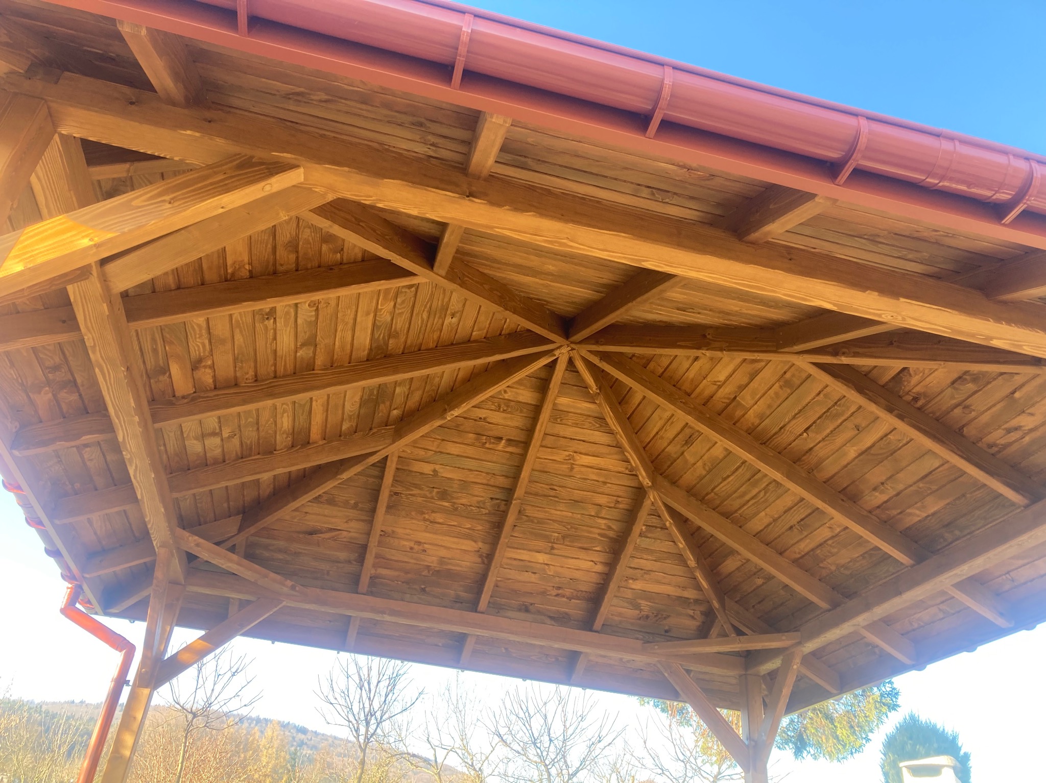 Drewniana konstrukcja zadaszenia w kształcie ośmiokąta, widok od dołu, z czerwonymi rynnami i elementami wykończeniowymi, na tle błękitnego nieba i drzew bez liści w oddali.