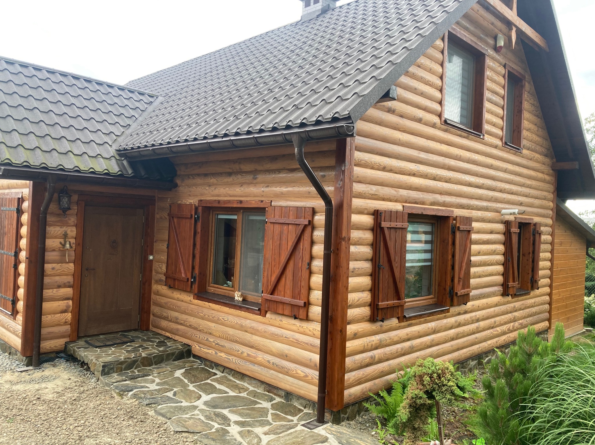Zewnętrzny widok domu z bali z drewnianymi okiennicami i kamiennymi schodami wejściowymi.