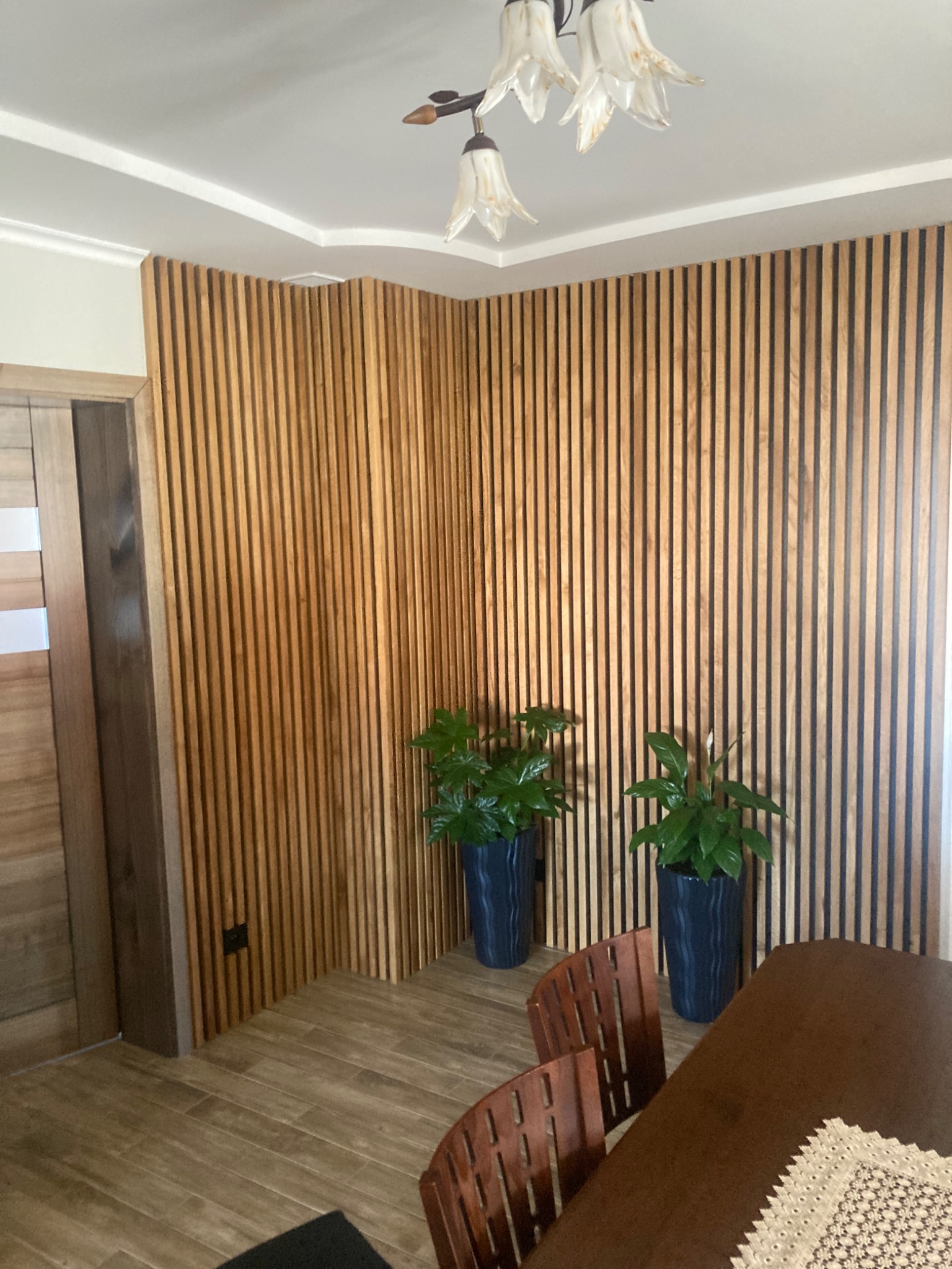 Drewniane lamele pionowe na ścianie w salonie, dwie donice z roślinami, widoczny fragment stołu i krzeseł oraz lampa sufitowa w kształcie kwiatów.