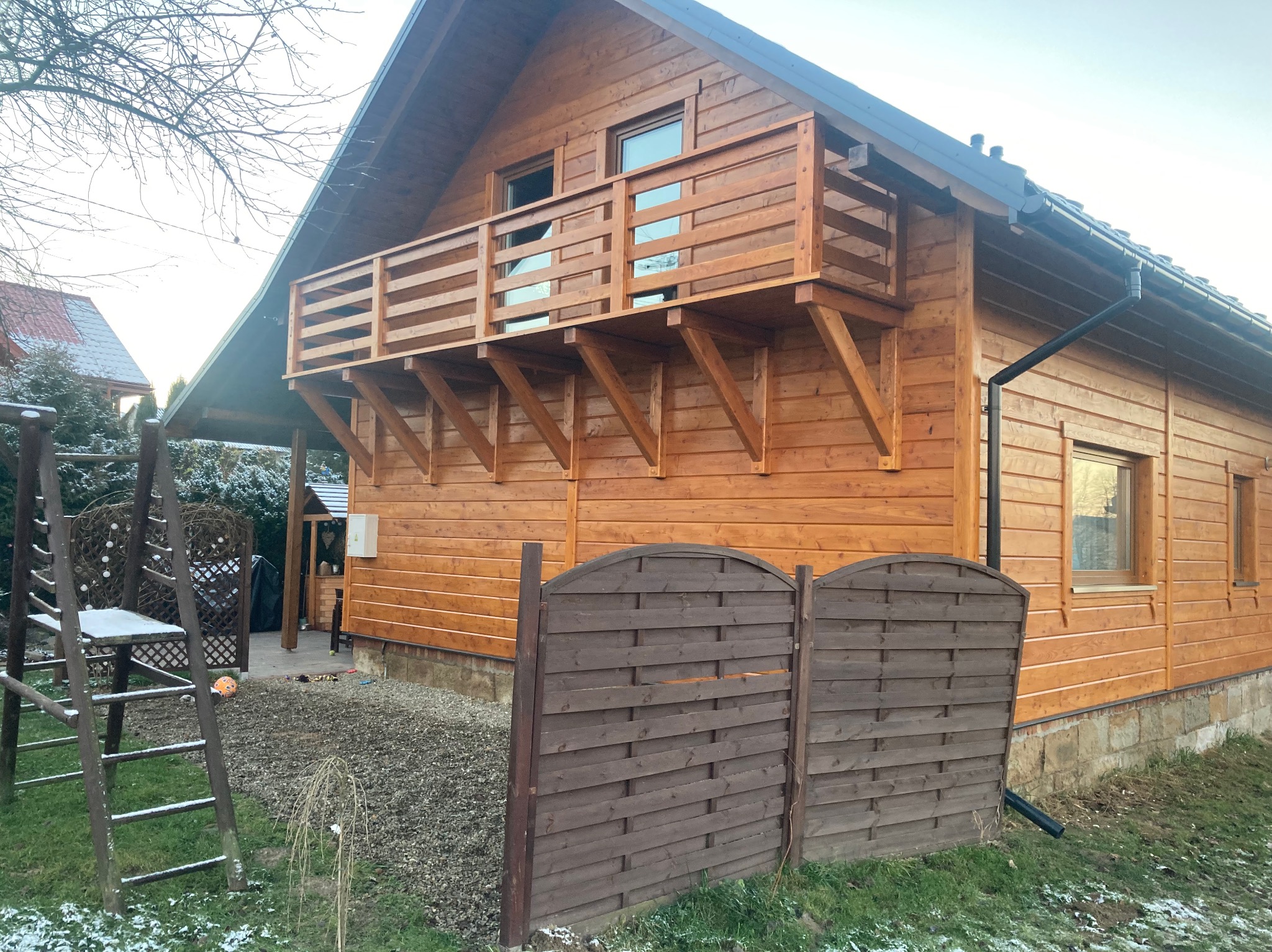 Drewniany domek z balkonem, widok z zewnątrz, częściowo ogrodzony brązowym płotem, na trawie leży śnieg.