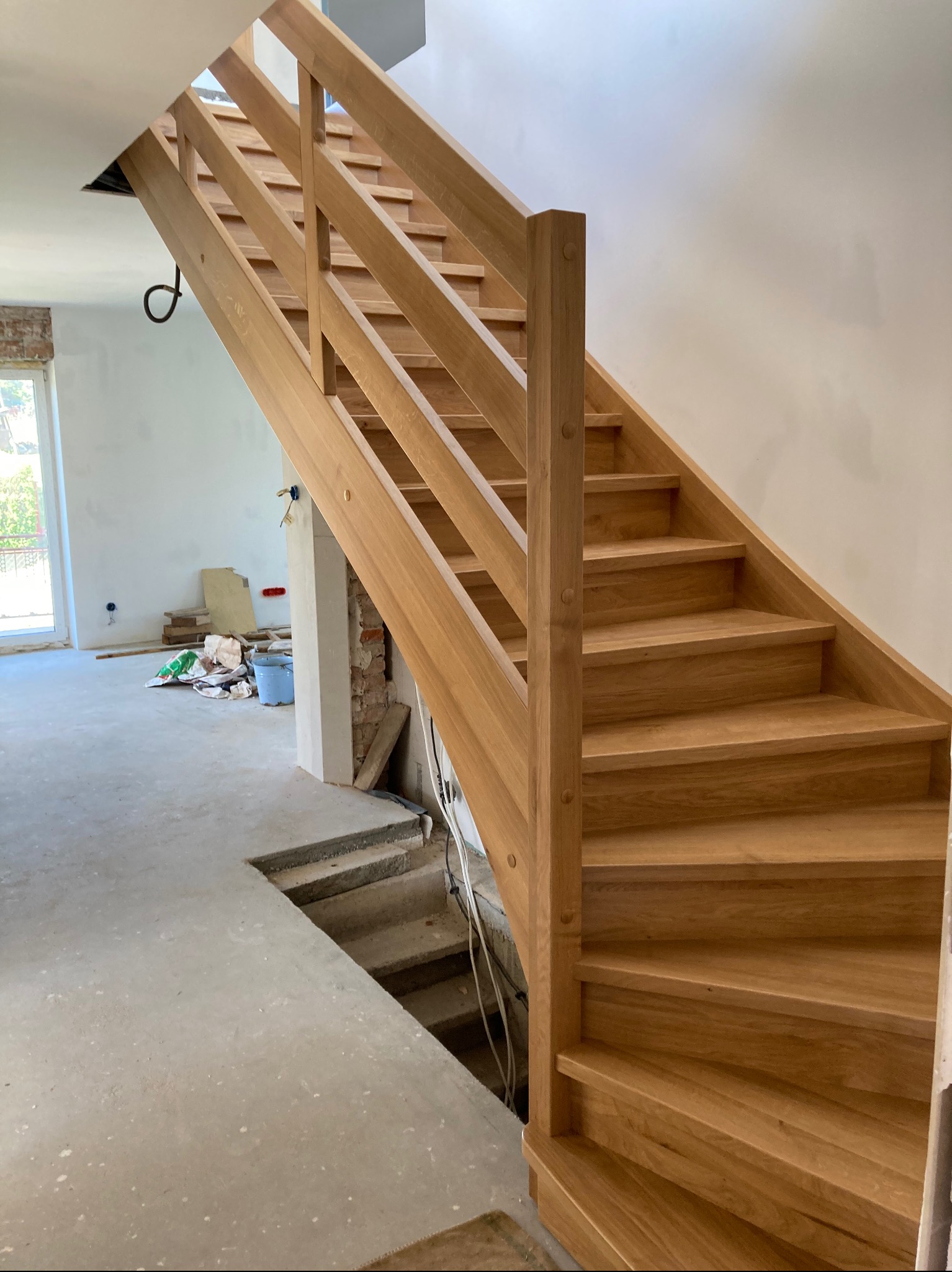 Jasne drewniane schody z balustradą, widoczne na tle surowego wnętrza w trakcie remontu, z betonową podłogą i fragmentami odsłoniętej cegły.