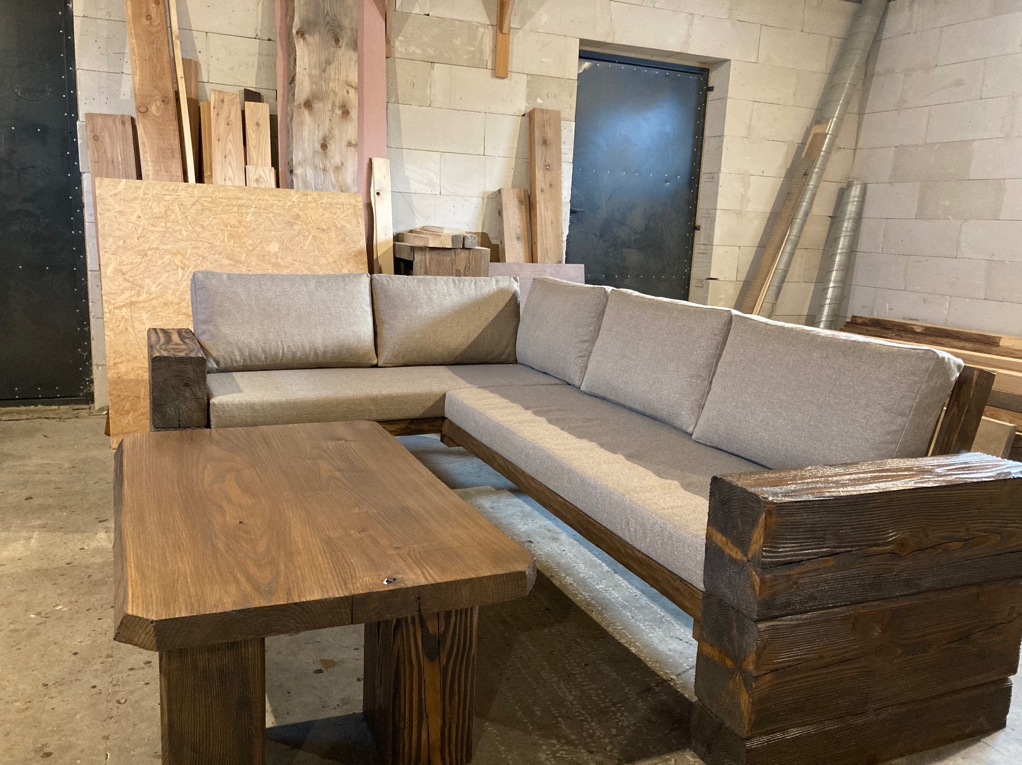 Kanciasty, drewniany stół i sofa narożna z jasną tapicerką, z grubych belek, w warsztacie z betonowych pustaków.