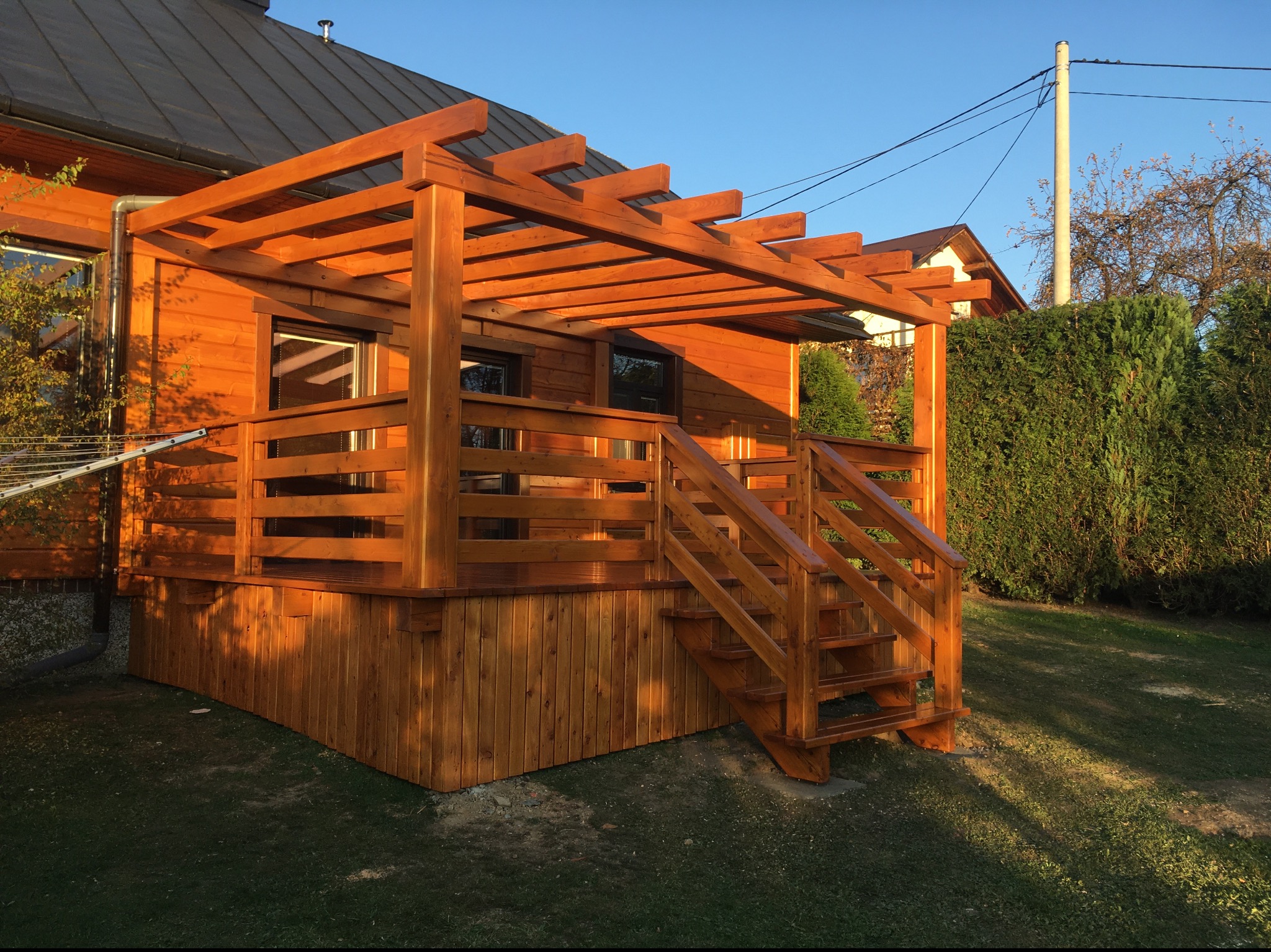 Drewniana konstrukcja tarasu z pergolą, schodami i balustradami, przylegająca do budynku mieszkalnego, oświetlona popołudniowym słońcem, z widocznymi elementami ogrodu w tle.