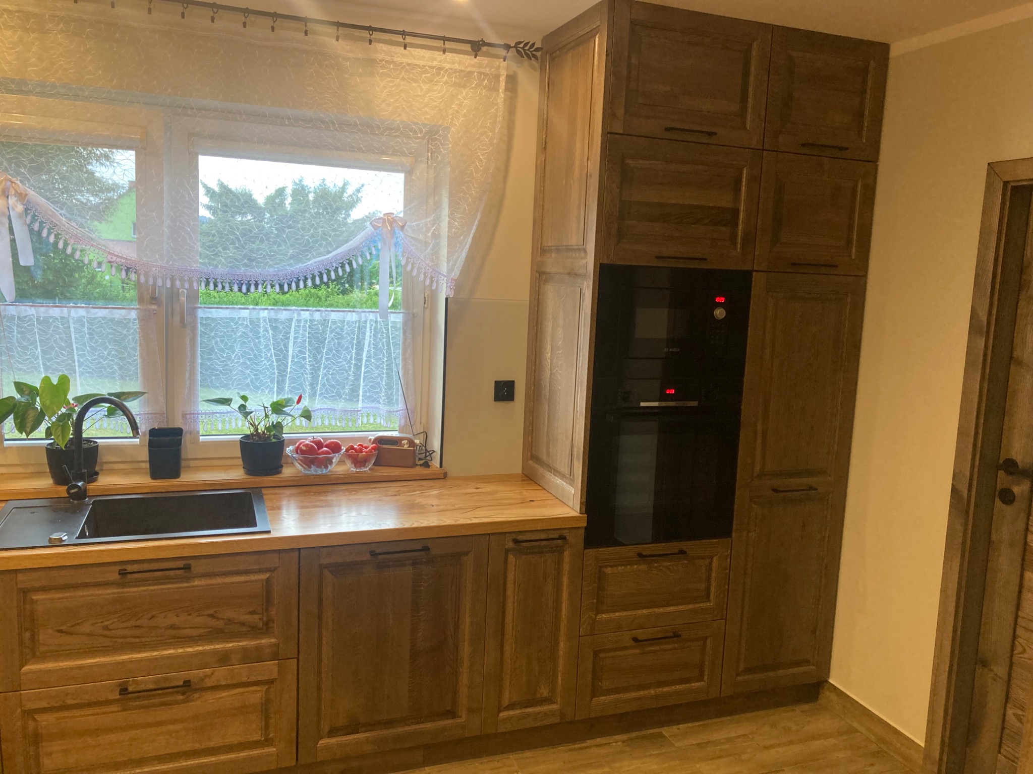 Kuchenna zabudowa z drewnianymi szafkami, blatem i wbudowanym piekarnikiem, z czarnym zlewem pod oknem ozdobionym firanką i roślinami.