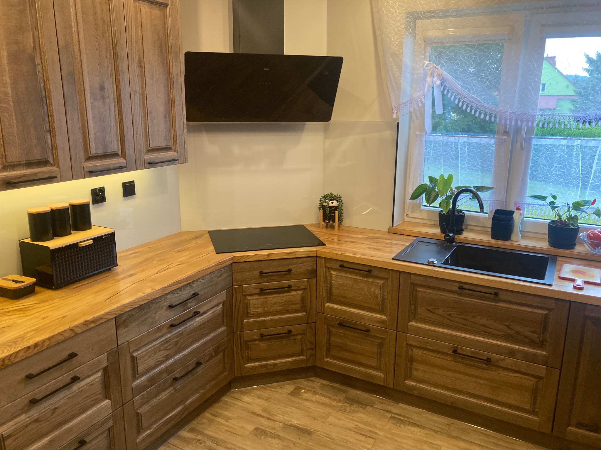 Drewniana zabudowa kuchenna z frezowanymi frontami, drewnianym blatem i czarną armaturą, widok na okno z firanką i roślinami.