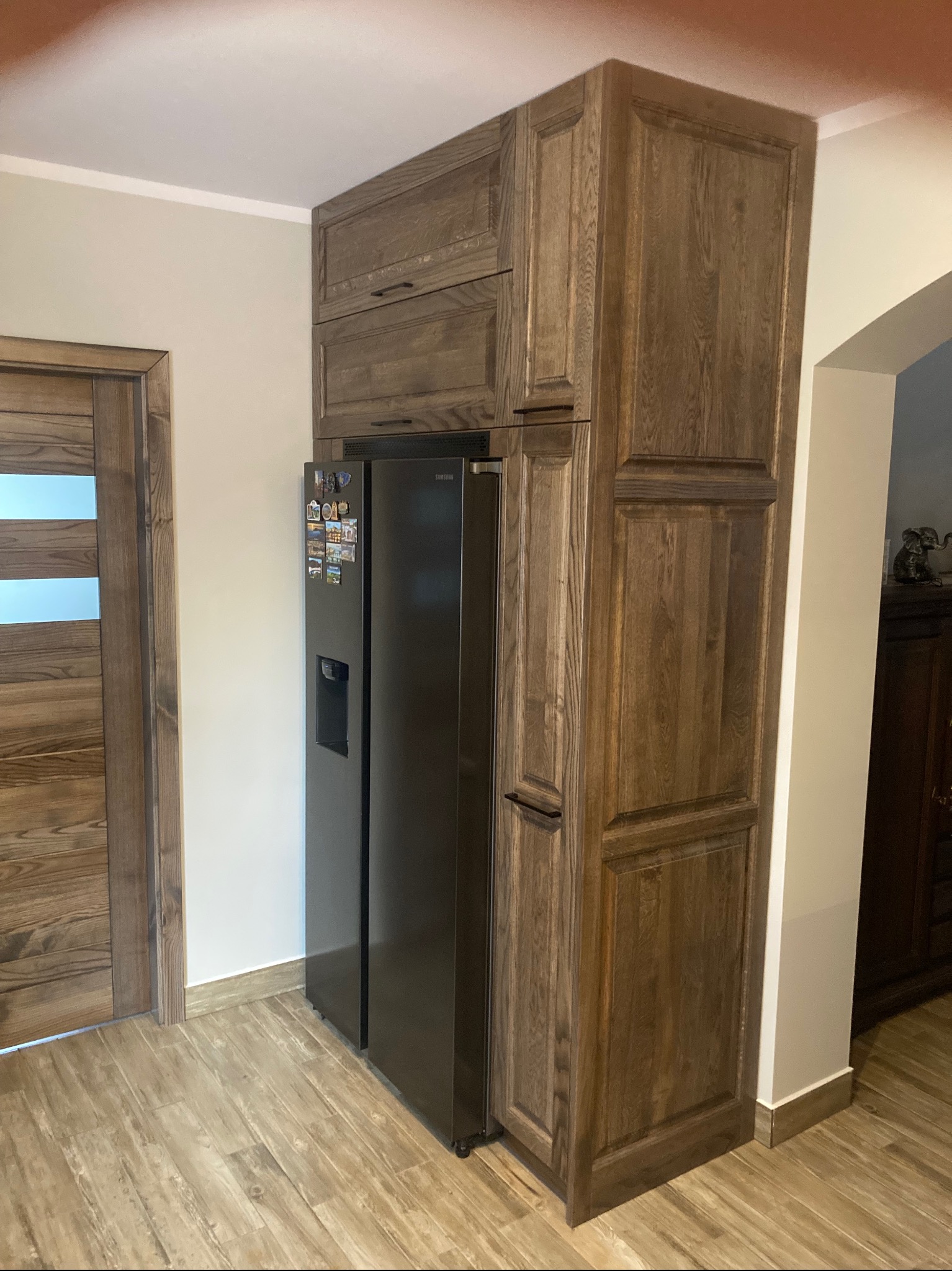 Drewniana zabudowa lodówki w kuchni z drewnianymi drzwiami w tle, widoczne magnesy na lodówce.