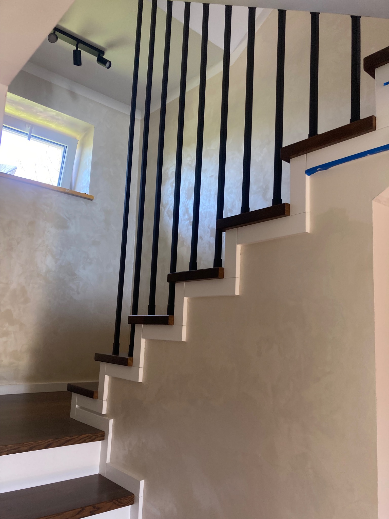 Nowoczesne schody z ciemnego drewna z czarnymi, metalowymi balustradami, białe stopnice, ściana wykończona dekoracyjnym tynkiem.