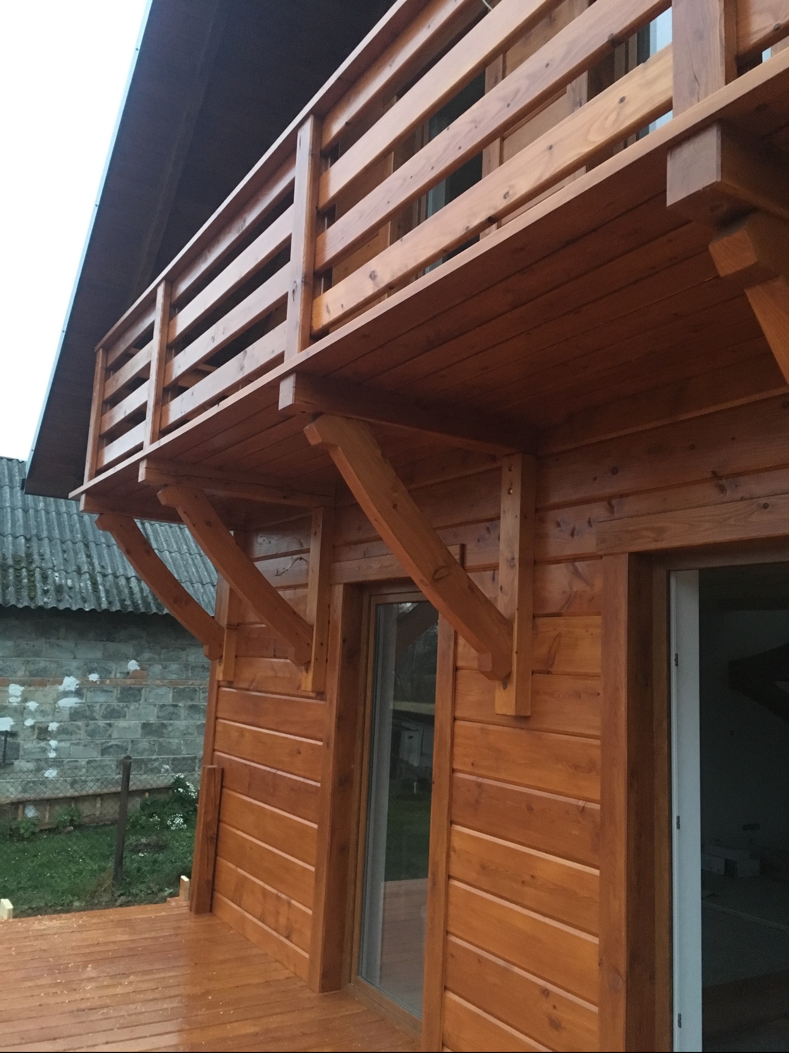 Zewnętrzna, drewniana balustrada balkonu z widocznymi wspornikami, wykonana z desek o ciepłym, brązowym odcieniu, przylegająca do ściany budynku z widocznymi drzwiami.