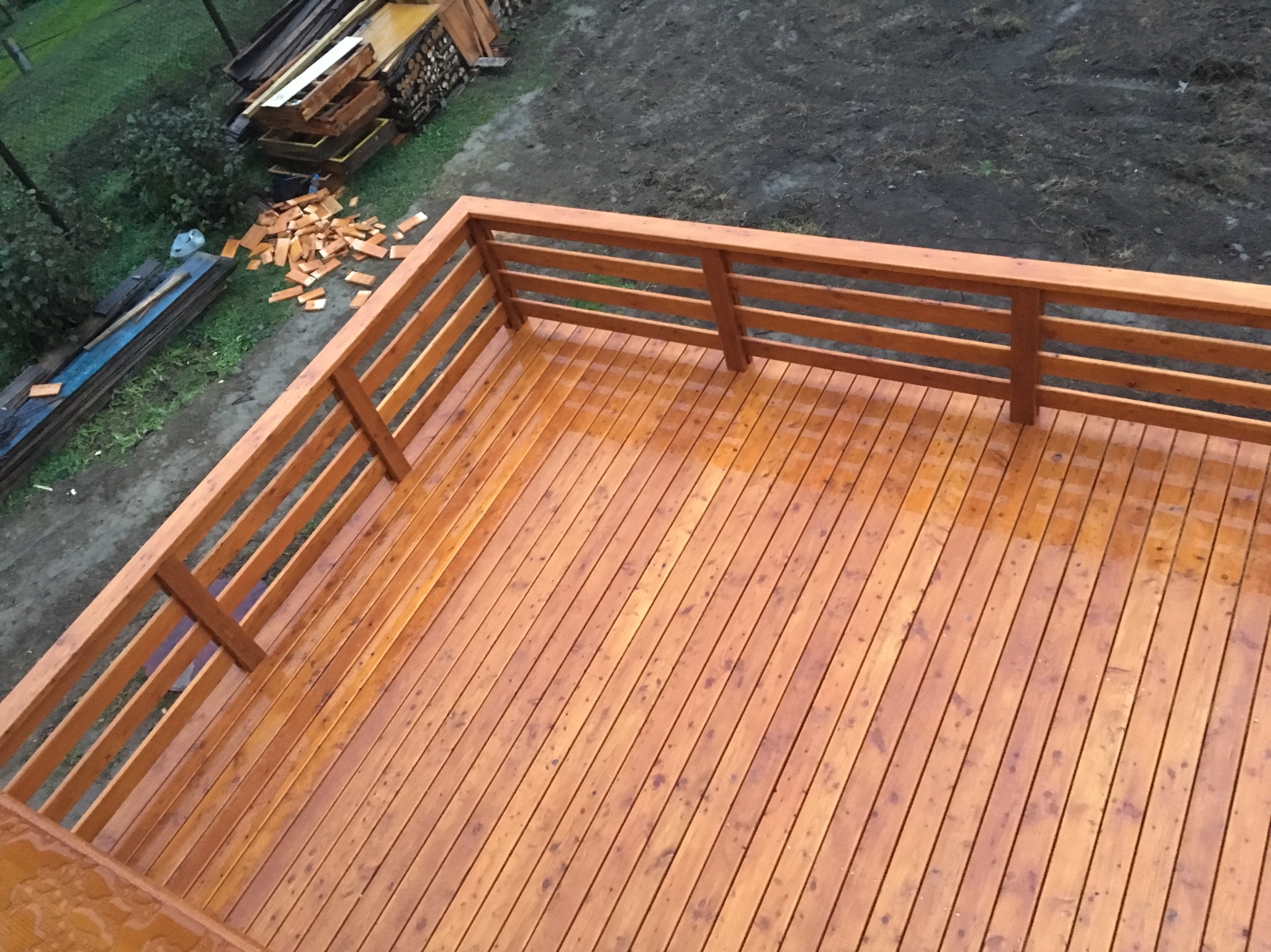Nowa, drewniana konstrukcja tarasu z balustradą, widok z góry na mokre deski po deszczu, w tle deski i ziemia.