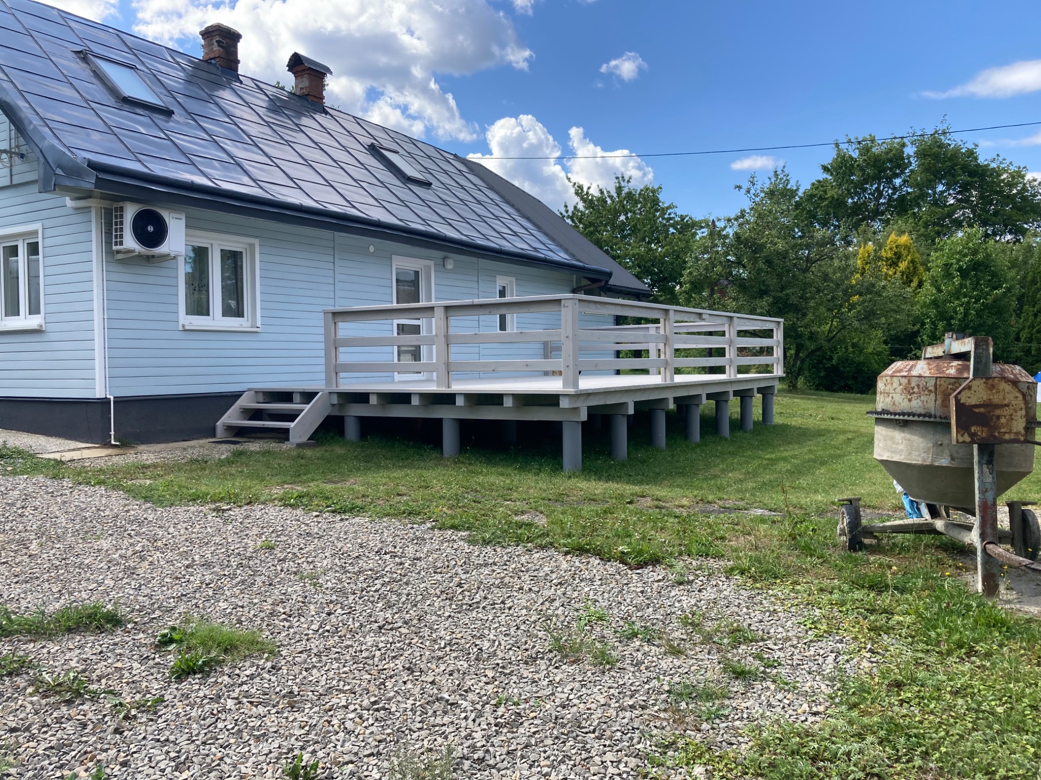 Jasny dom z nowym, podwyższonym tarasem z drewnianą balustradą. Widoczne schody wejściowe i betonowa mieszarka. Błękitne niebo z obłokami w tle.