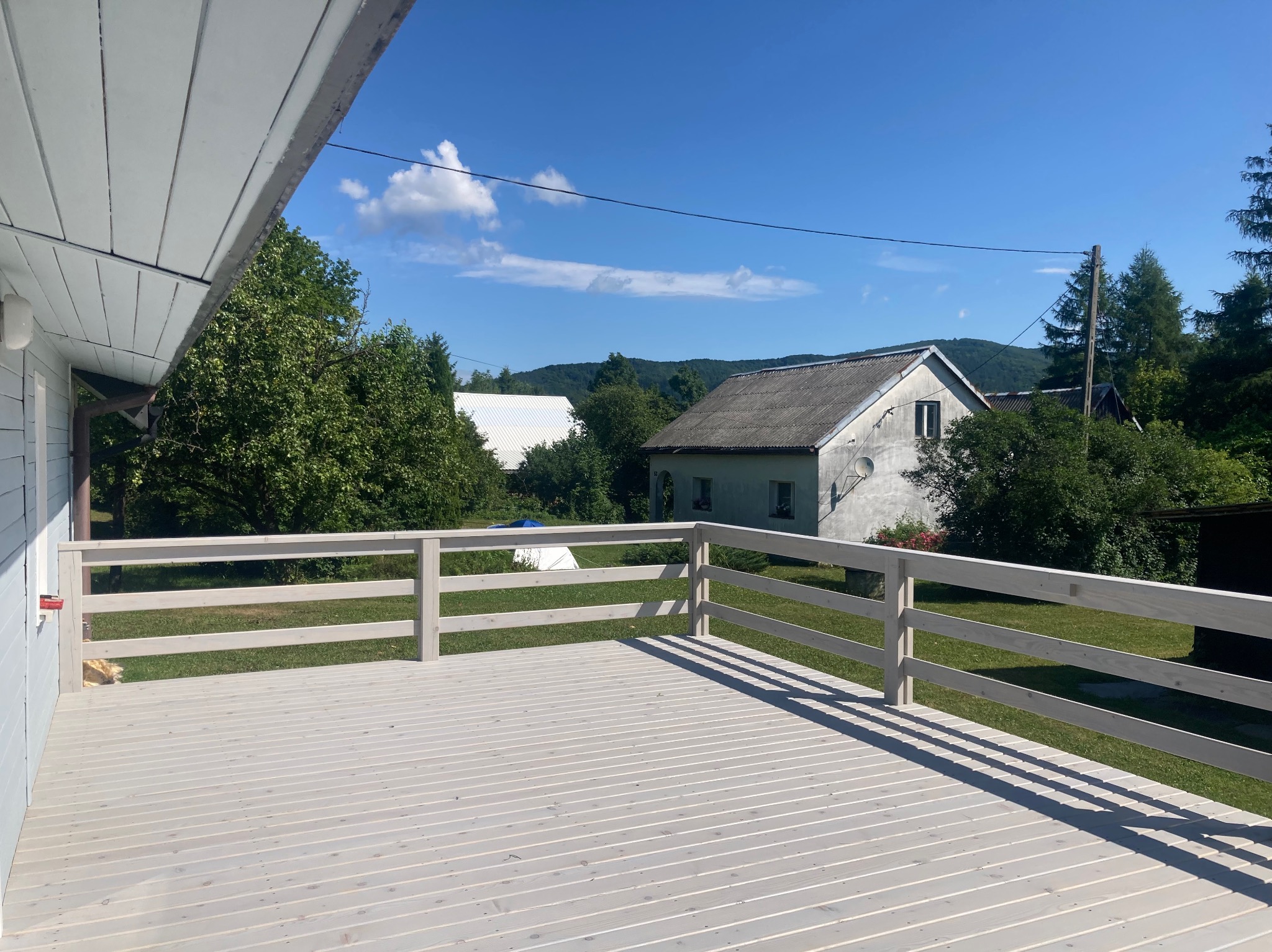 Jasny, drewniany taras z barierkami w odcieniach szarości, widok na zielony ogród i dom w tle, słoneczny dzień, cień barierki na deskach.