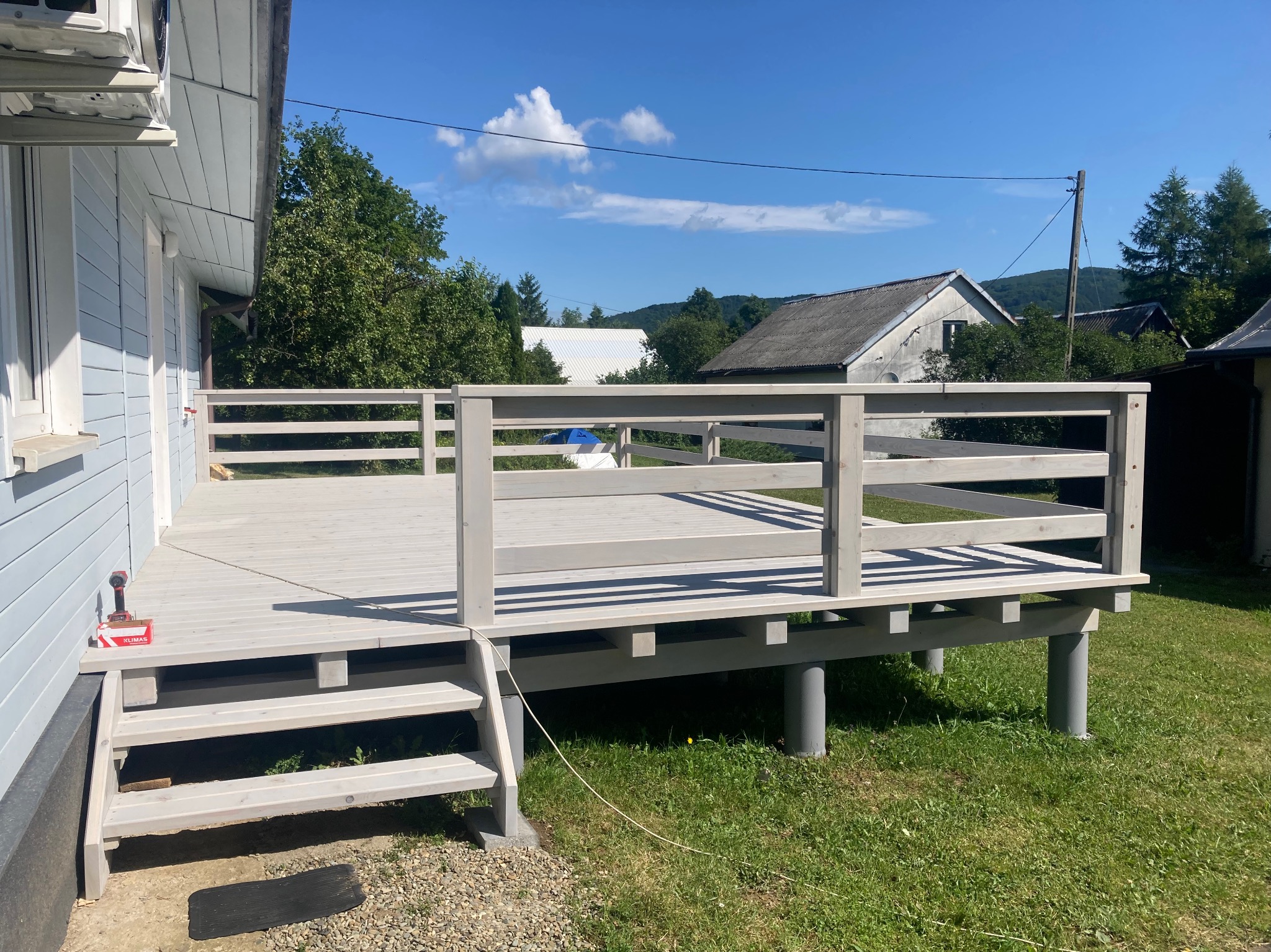 Jasnoszary taras z balustradą, widok na ogród i domy w tle. Schody prowadzą na taras. W rogu wkrętarka i pudełko z wkrętami.