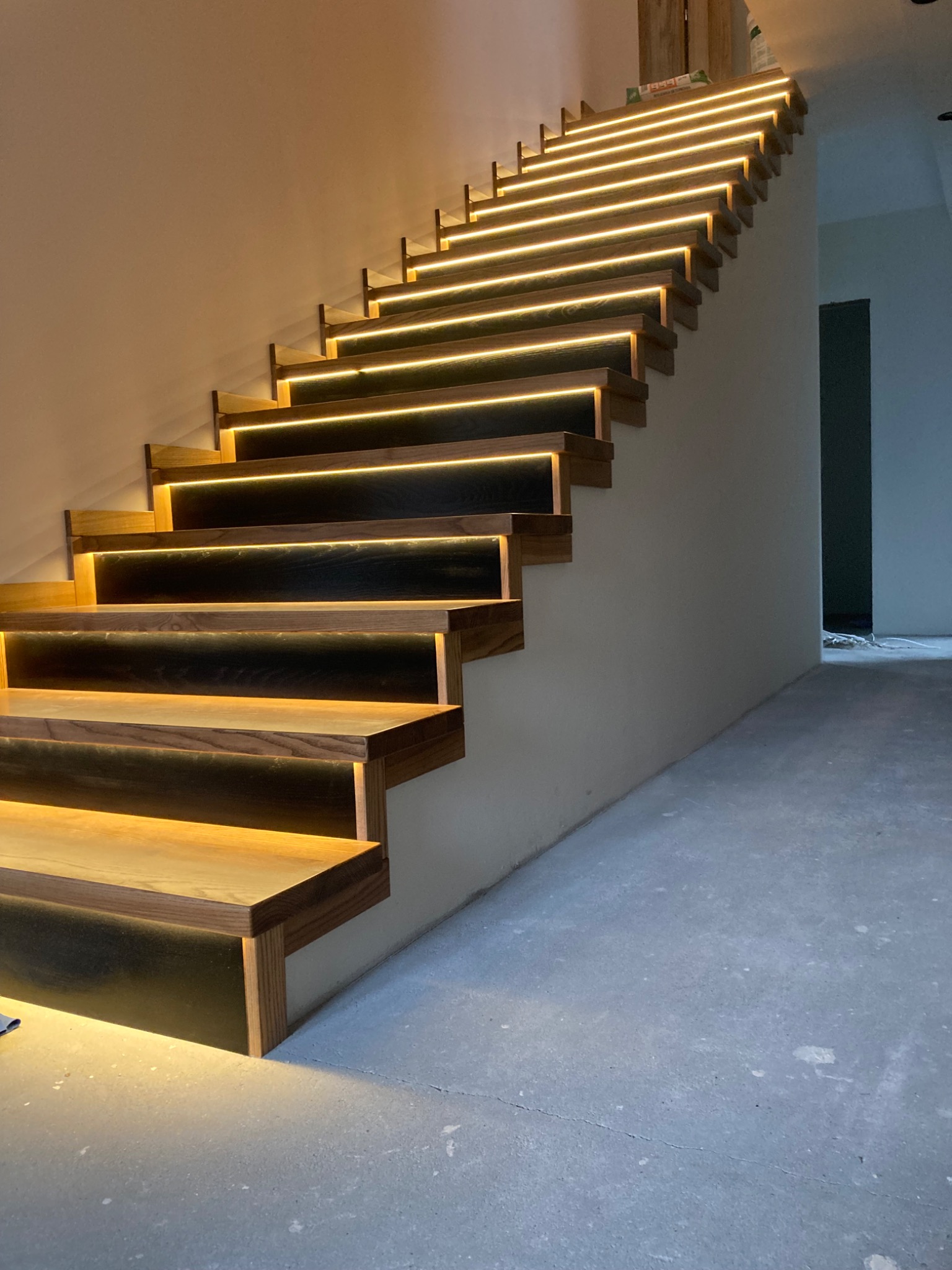 Nowoczesne schody z drewna z podświetleniem LED, widok z dołu, ukazujący detale wykończenia i ciepłe światło oświetlające betonową podłogę.
