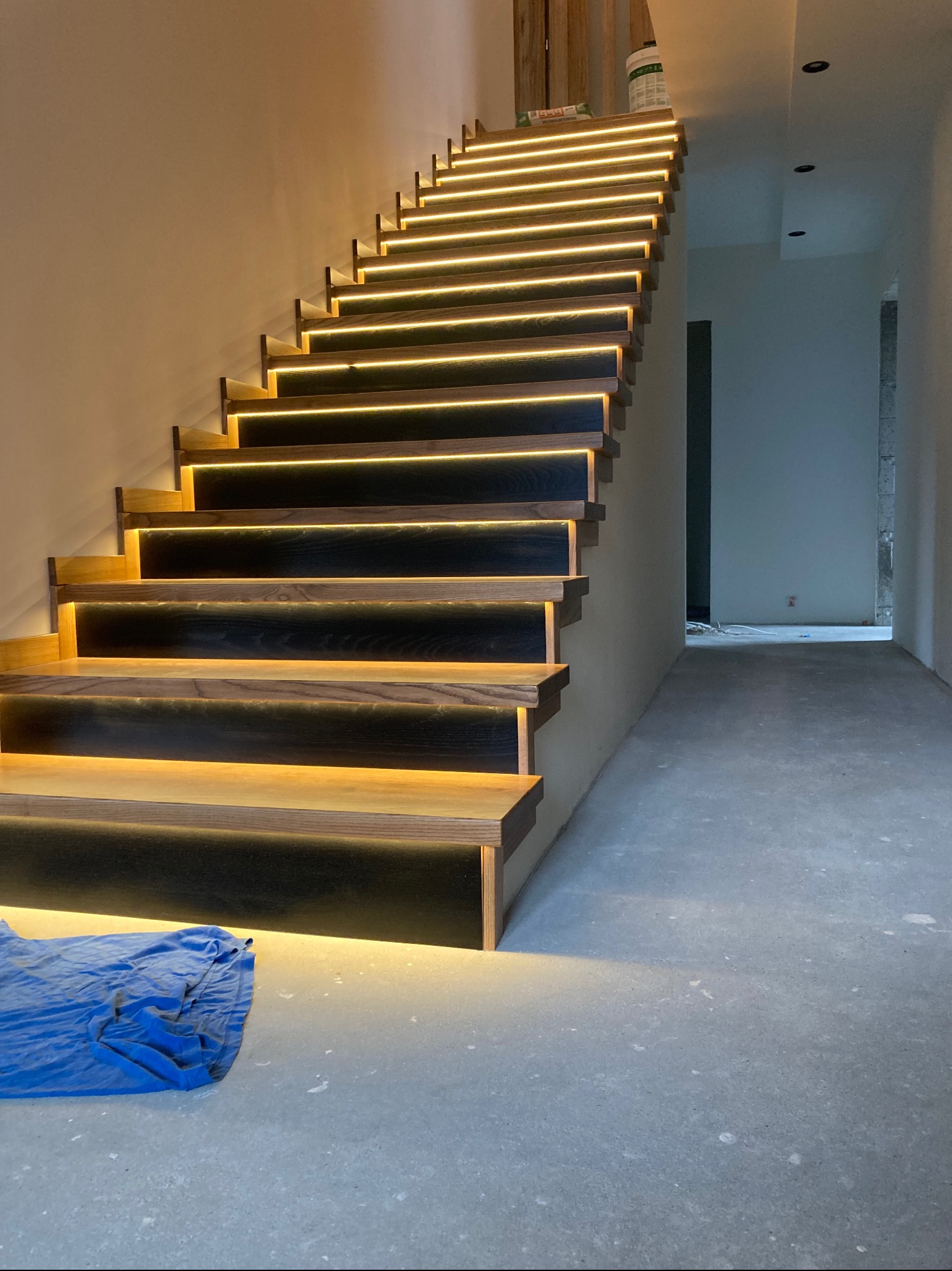 Nowoczesne schody z ciemnego drewna z subtelnym, ciepłym oświetleniem LED pod każdym stopniem. Na podłodze leży niebieska plandeka. W tle surowy stan wykończenia.