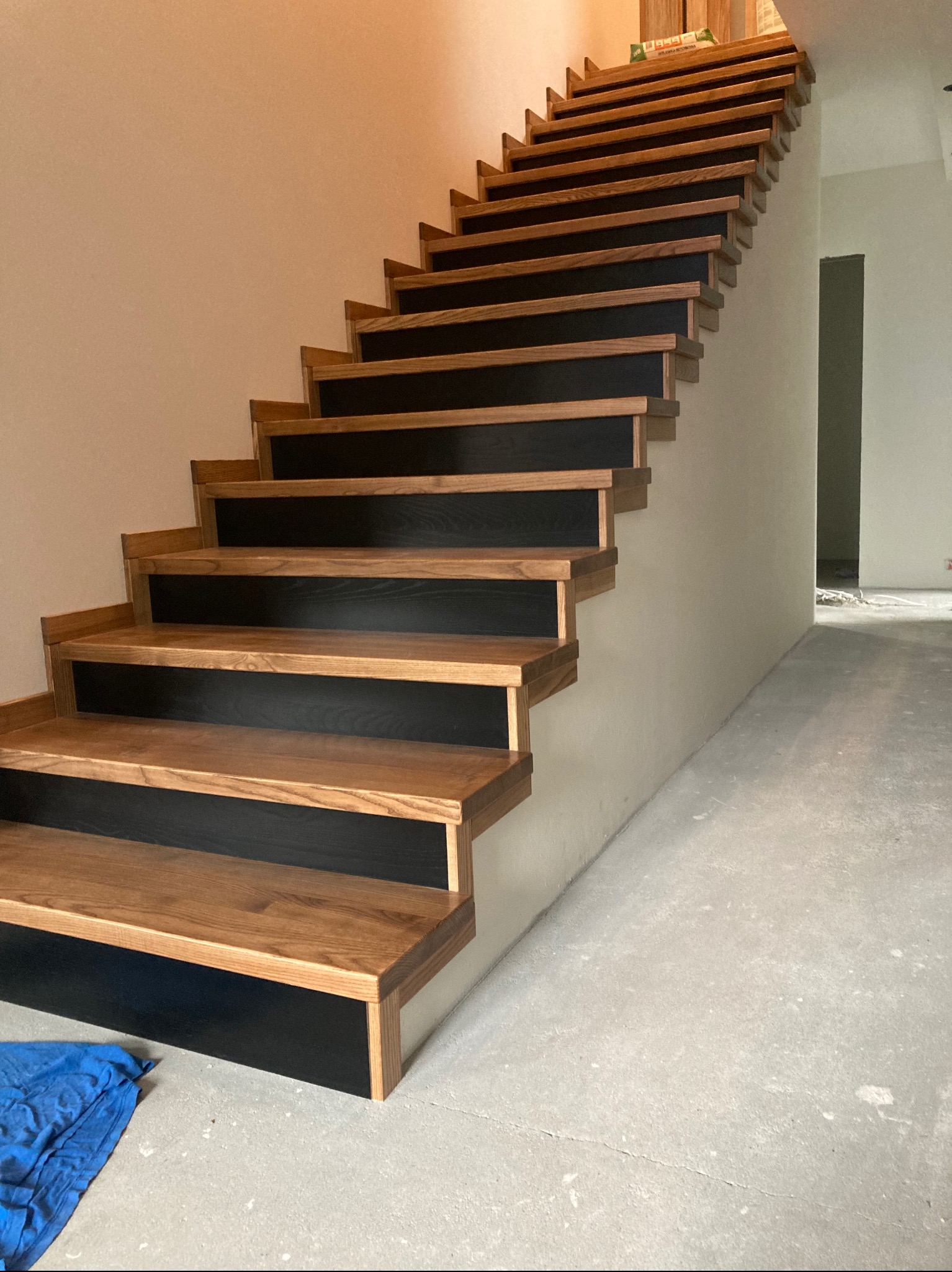 Nowoczesne schody z drewnianymi stopniami i czarnymi podstopnicami w surowym wnętrzu. Widoczne niedoskonałości podłoża i brak wykończenia ścian.