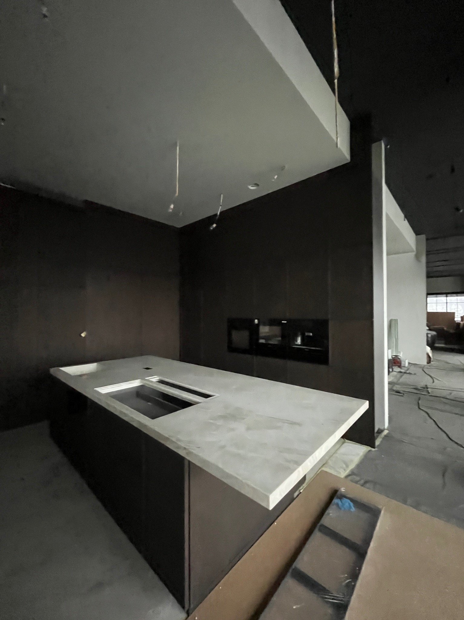 Surowa kuchnia w trakcie wykończenia: wyspa z jasnym blatem i ciemnymi szafkami, w tle zabudowa z piekarnikiem i mikrofalówką, widoczne przewody i materiały budowlane.