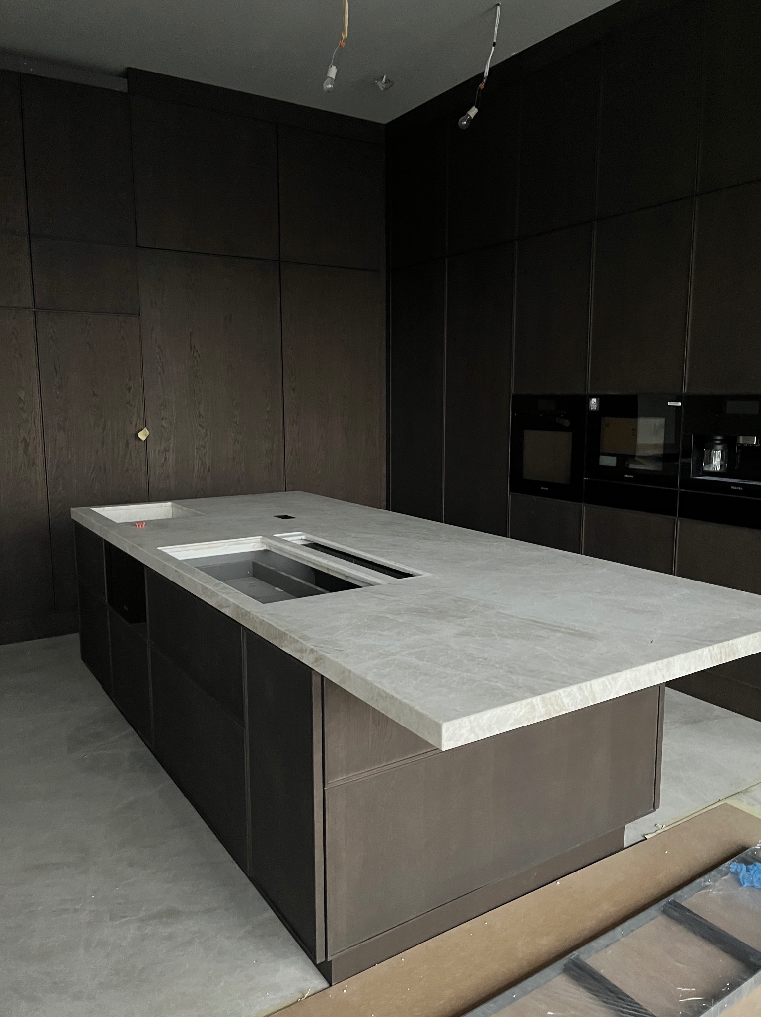 Nowoczesna kuchnia w trakcie wykańczania: ciemne, drewniane szafki z wbudowanymi urządzeniami AGD, jasny blat wyspy z przygotowanymi otworami na zlew i inne akcesoria, surowy beton na podłodze...