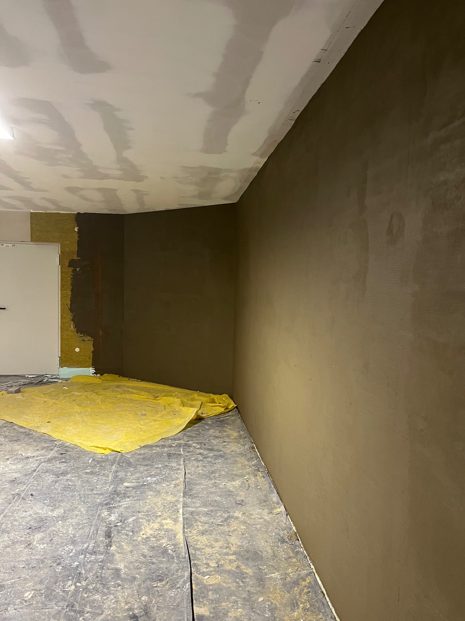Wnętrze w trakcie prac wykończeniowych: ściana pokryta świeżą warstwą gładzi w odcieniach brązu, widoczna izolacja ściany z wełny mineralnej, podłoga zabezpieczona żółtą folią malarską, sufit...