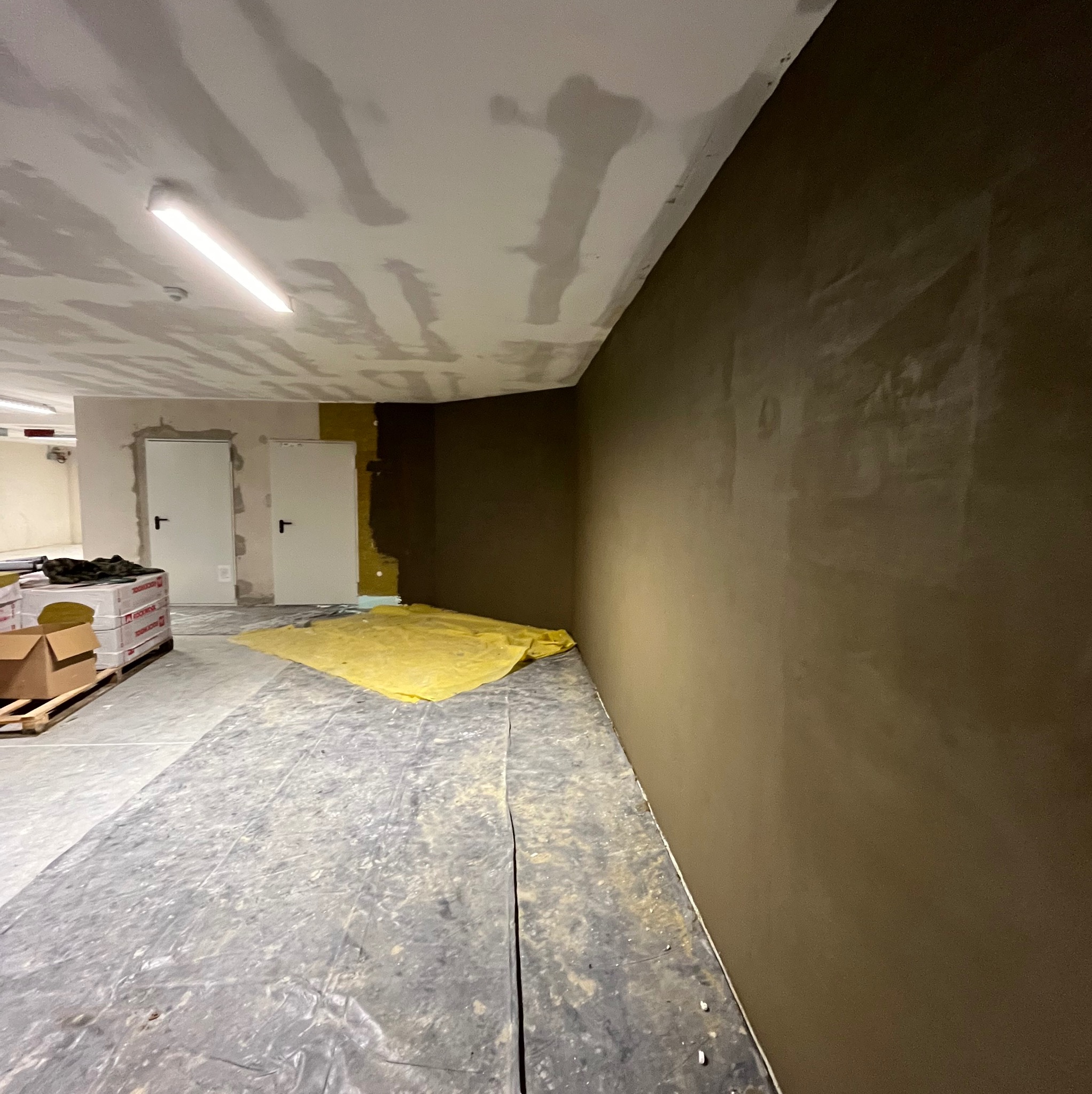 Przygotowana ściana w odcieniach brązu, częściowo pokryta gładzią, podłoga zabezpieczona folią, widoczne materiały budowlane na palecie i dwie białe drzwi w tle.