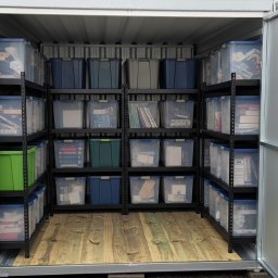 Miejski Box - Wnętrze kontenera magazynowego z otwartymi drzwiami, wyposażonego w regały z plastikowymi pojemnikami. Podłoga z desek, metalowe ściany kontenera.