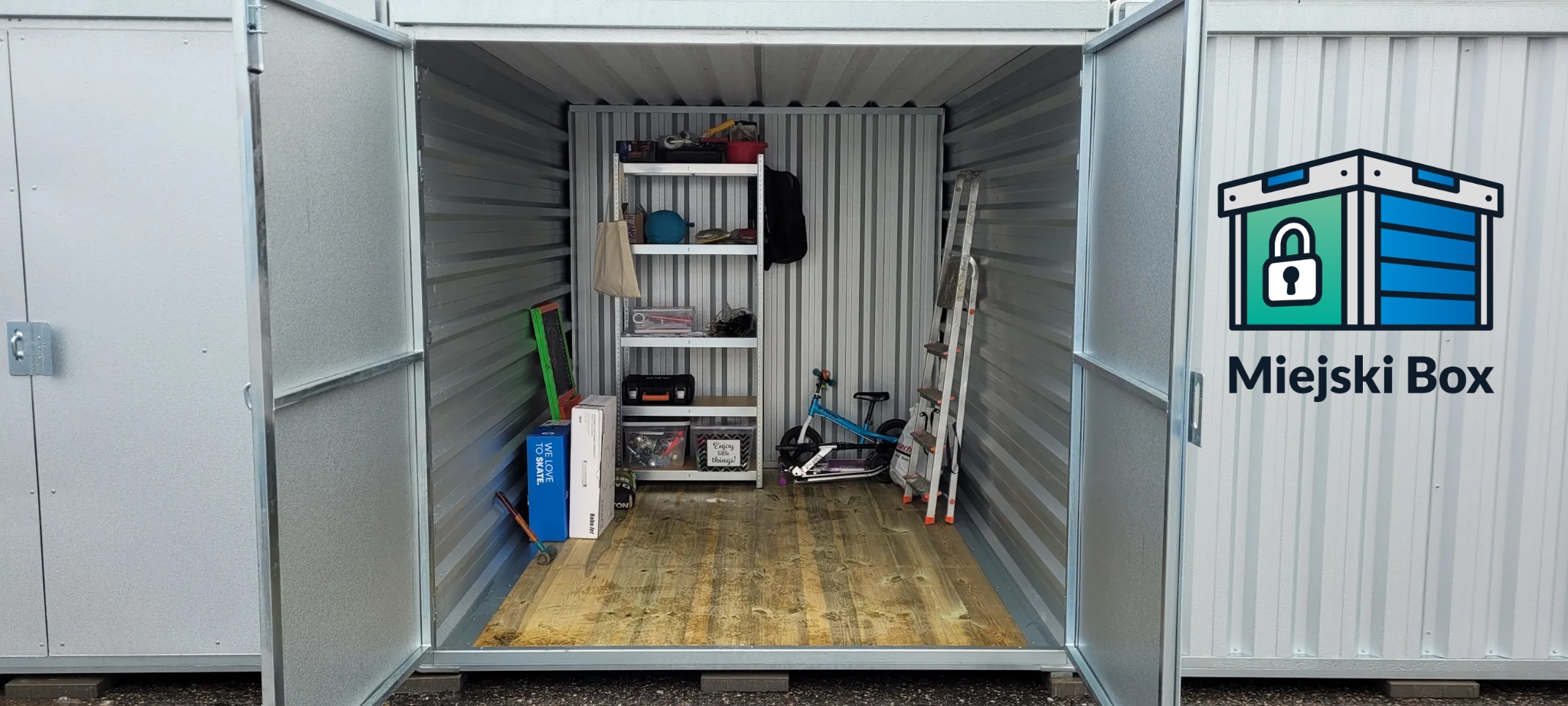 Otwarty, metalowy kontener magazynowy z drewnianą podłogą. W środku regał, drabina, rower i kartony. Widok z zewnątrz, otwarte drzwi.