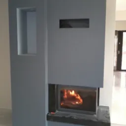 Szary kominek obudowany płytami kartonowo-gipsowymi z widocznym płomieniem, wnęką i prostokątnym otworem wentylacyjnym, w trakcie wykańczania.