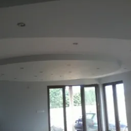 Nowoczesny salon z podwieszonym sufitem o okrągłym kształcie i punktowym oświetleniem, widoczne okna balkonowe i fragment ściany z dekoracyjną fototapetą