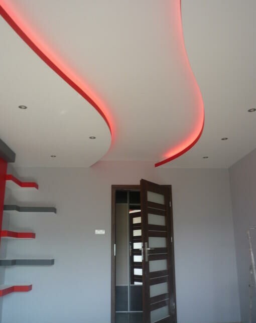 Nowoczesne wnętrze z podwieszanym sufitem o falistym kształcie z czerwonym oświetleniem LED, widoczne drzwi z przeszkleniami i półki na ścianie w kolorach szaro-czerwonych.