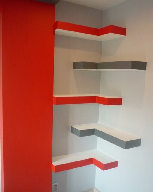 Narożne, asymetryczne półki ścienne w kolorach czerwonym, białym i szarym, zamontowane na białej ścianie, widoczny fragment czerwonej ściany i gniazdko elektryczne.