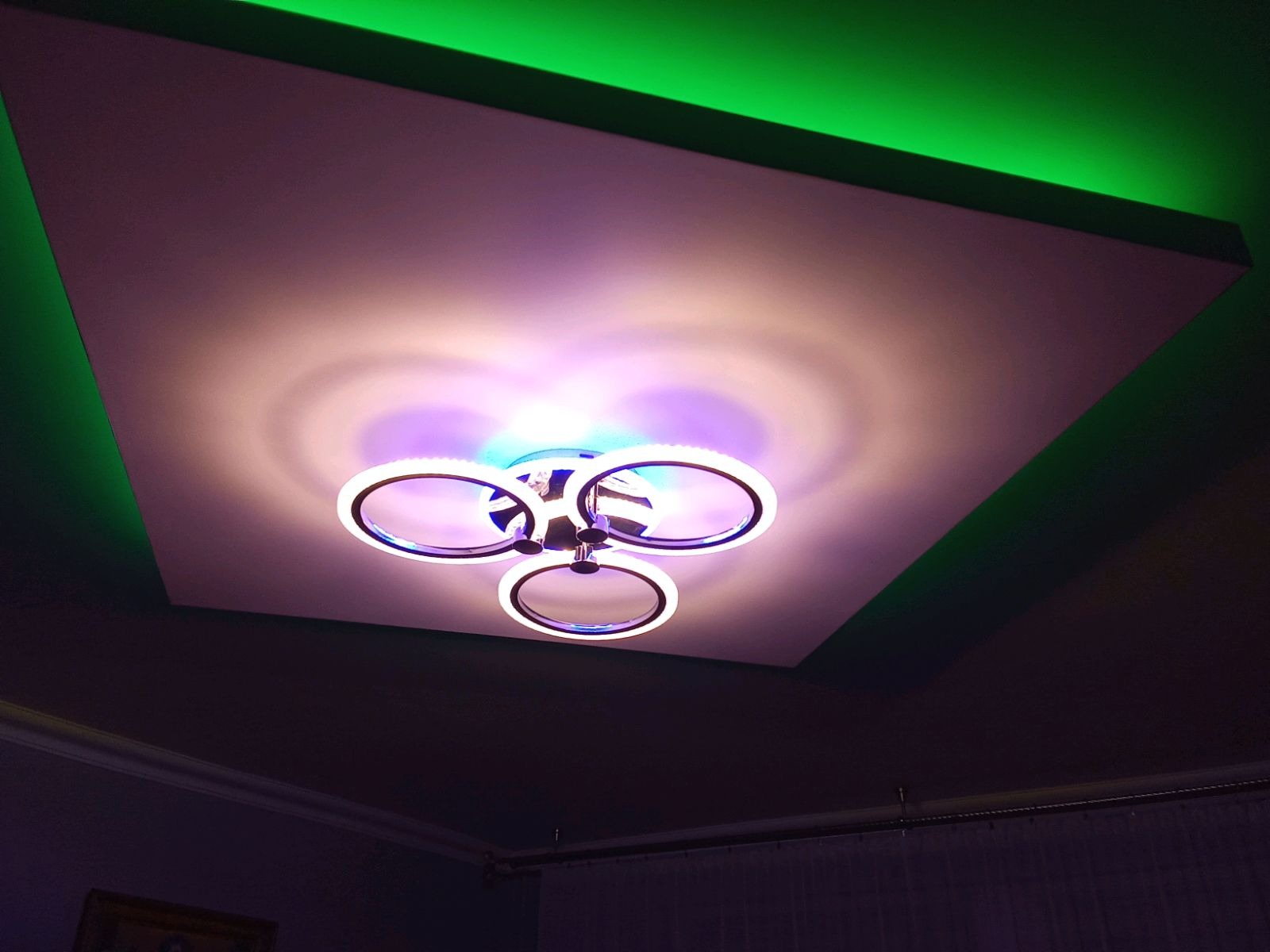 Sufit podwieszany z oświetleniem LED w kolorze zielonym i nowoczesną lampą sufitową z trzema okręgami, oświetlającą pomieszczenie fioletowo-białym światłem.