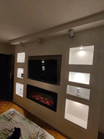 Ściana z wnękami podświetlonymi punktowym oświetleniem, w centralnej części telewizor i elektryczny kominek, widoczne elementy dekoracyjne w niszach.