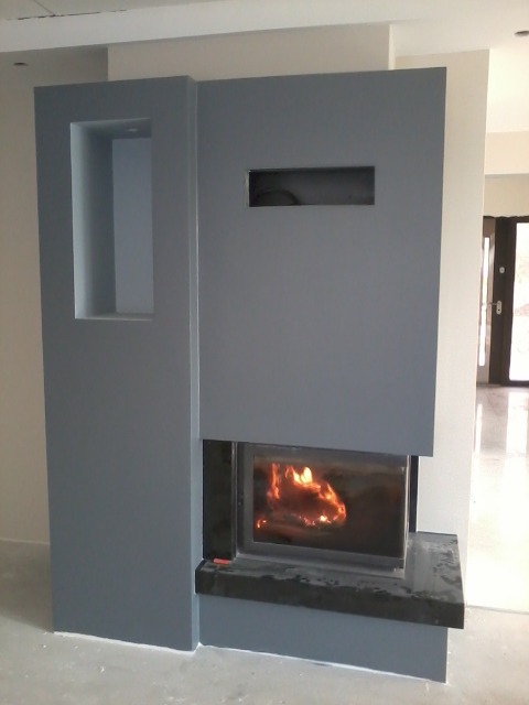 Szary kominek obudowany płytami kartonowo-gipsowymi z widocznym płomieniem, wnęką i prostokątnym otworem wentylacyjnym, w trakcie wykańczania.