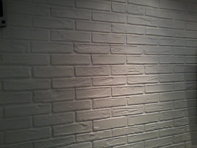Ściana wykończona białymi cegłami dekoracyjnymi, oświetlona z góry, z widocznym fragmentem instalacji elektrycznej w lewym górnym rogu.