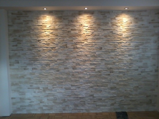Ściana wykończona dekoracyjną okładziną kamienną w odcieniach beżu i szarości, oświetlona trzema punktowymi lampami sufitowymi, z widoczną podłogą z paneli w ciepłym odcieniu drewna.