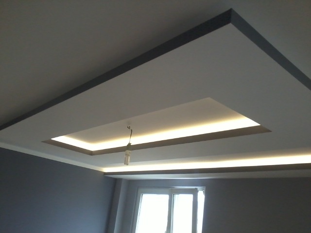 Podwieszany sufit z oświetleniem LED, wnęka w kształcie prostokąta z dodatkowym oświetleniem, szare ściany, widoczna żarówka na kablu, okno w tle.