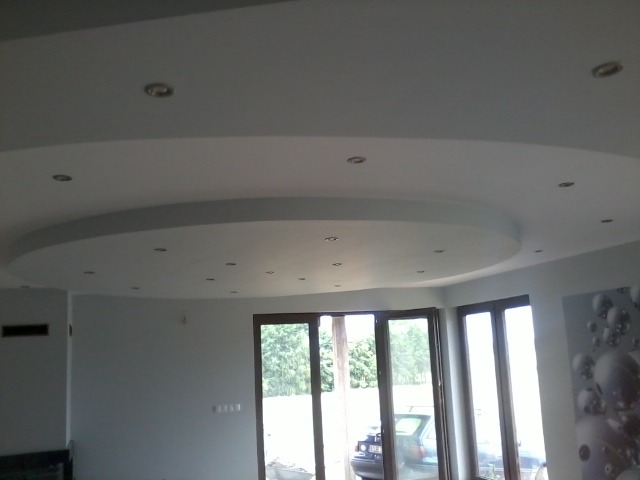 Nowoczesny salon z podwieszonym sufitem o okrągłym kształcie i punktowym oświetleniem, widoczne okna balkonowe i fragment ściany z dekoracyjną fototapetą