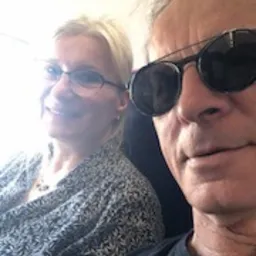 Selfie dwóch osób w samolocie: mężczyzna w czarnych okularach przeciwsłonecznych i czarnym T-shircie na pierwszym planie, uśmiechnięta kobieta w okularach i wzorzystej bluzce w tle, widok z okna...