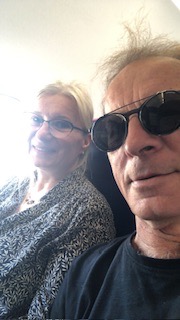 Selfie dwóch osób w samolocie: mężczyzna w czarnych okularach przeciwsłonecznych i czarnym T-shircie na pierwszym planie, uśmiechnięta kobieta w okularach i wzorzystej bluzce w tle, widok z okna...