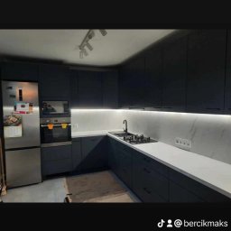 MAJSIER - Nowoczesna kuchnia w odcieniach grafitu z białym blatem, oświetleniem LED podszafkowym i srebrną lodówką. Na podłodze widoczny karton po transporcie.