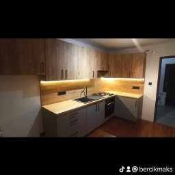 MAJSIER - Nowoczesna kuchnia w odcieniach drewna i szarości z podświetleniem LED pod szafkami wiszącymi, czarny zlew i płyta gazowa.
