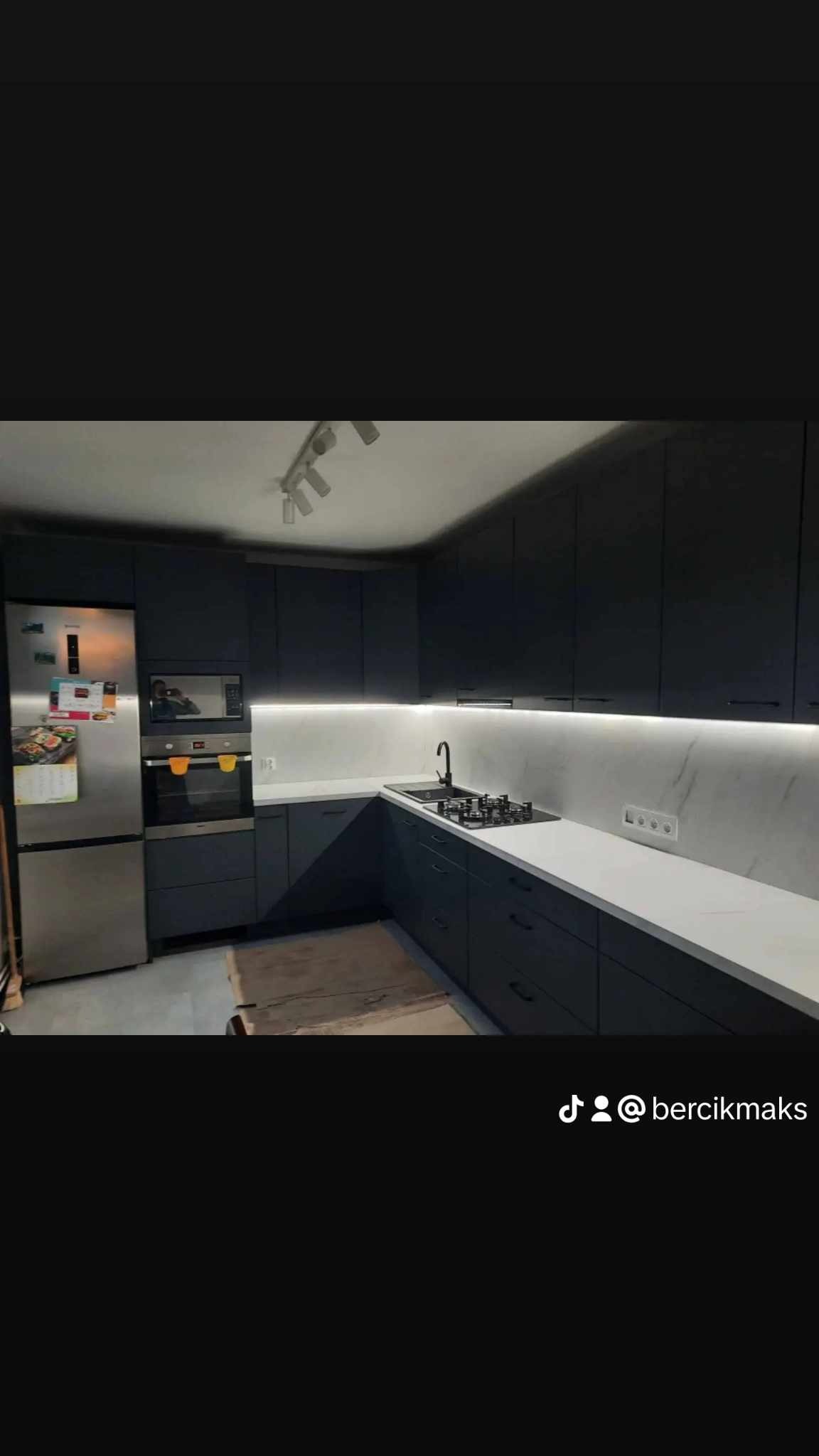 Nowoczesna kuchnia w odcieniach grafitu z białym blatem, oświetleniem LED podszafkowym i srebrną lodówką. Na podłodze widoczny karton po transporcie.