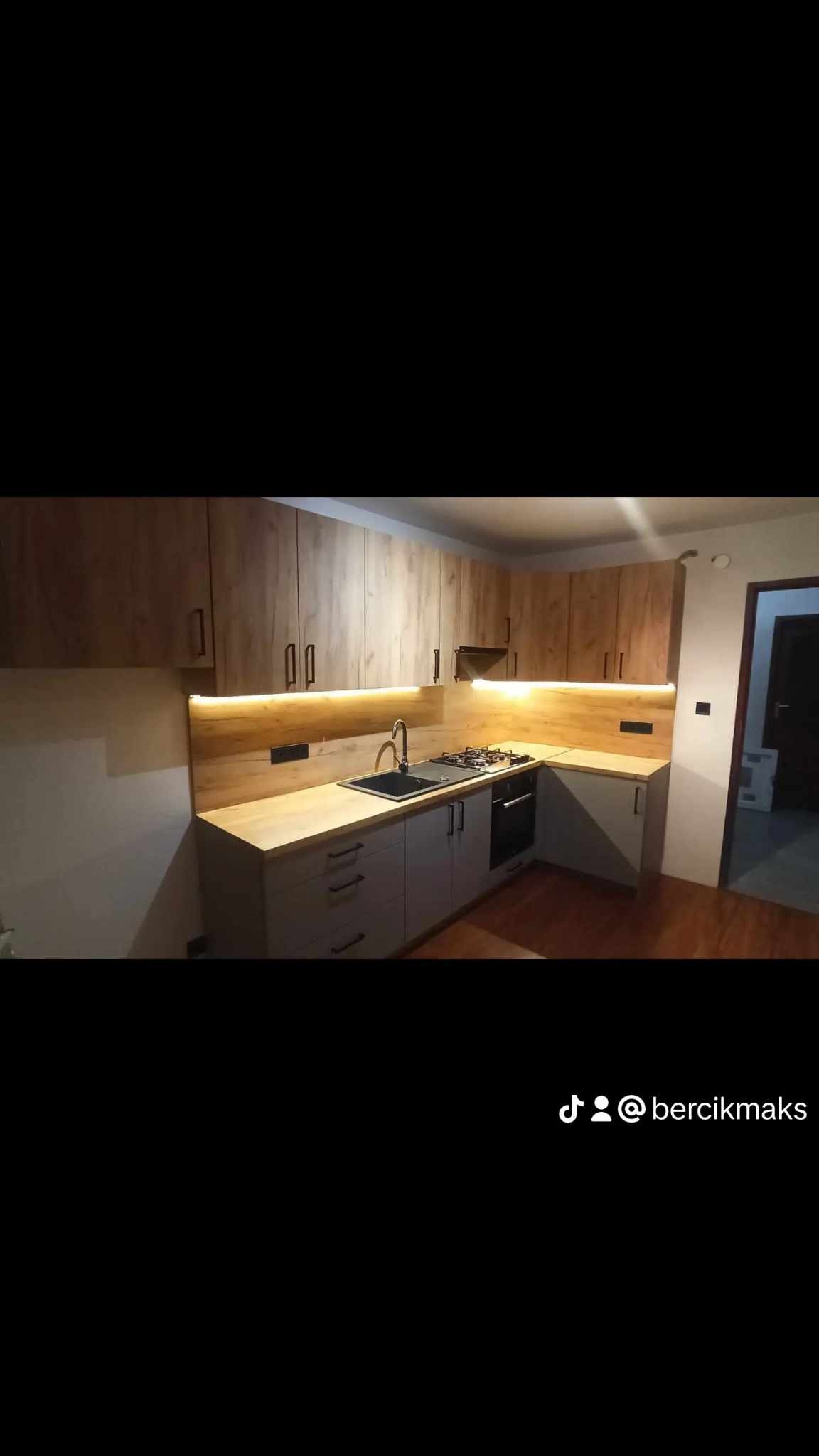 Nowoczesna kuchnia w odcieniach drewna i szarości z podświetleniem LED pod szafkami wiszącymi, czarny zlew i płyta gazowa.