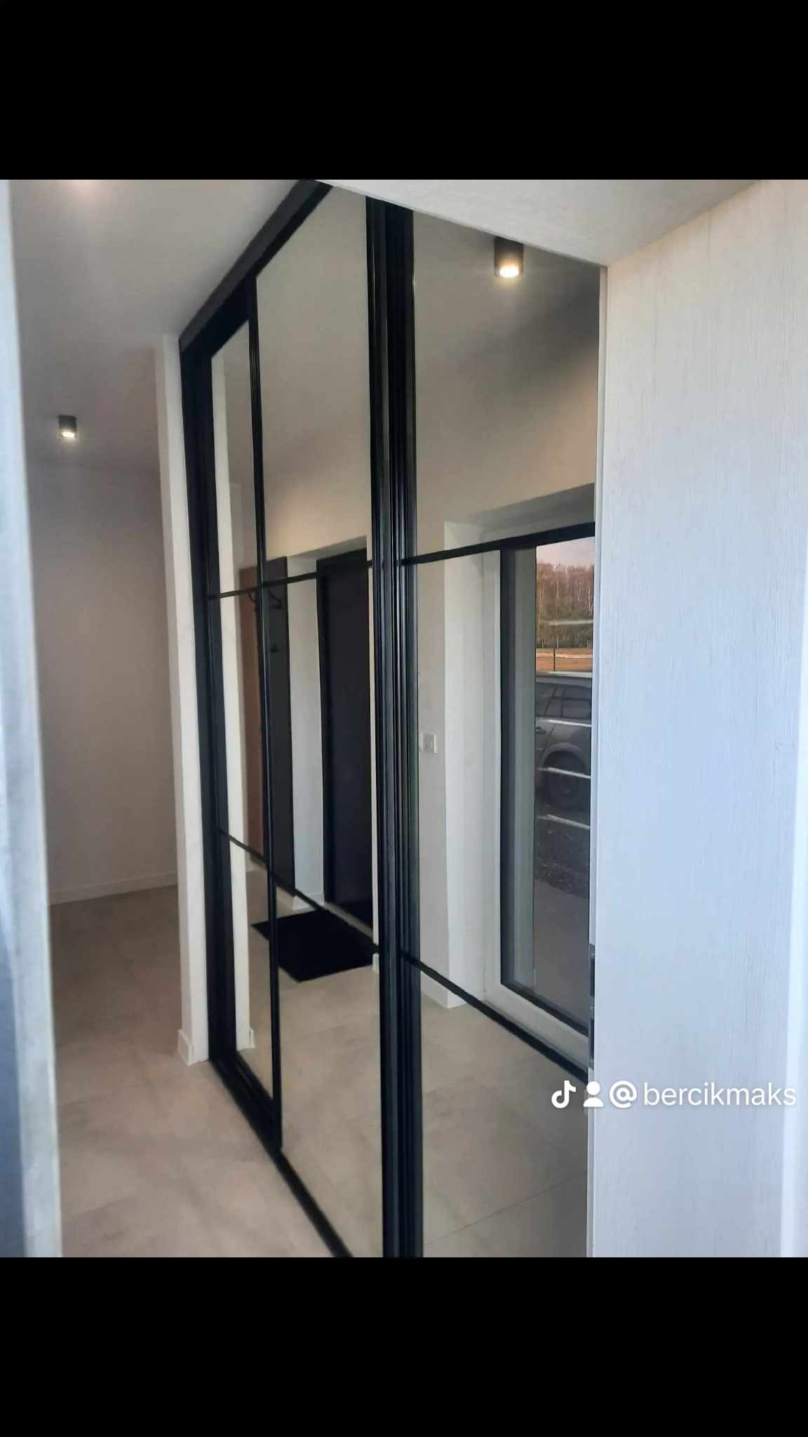 Przesuwne drzwi w stylu loft z czarnymi szprosami, oddzielające korytarz z jasną podłogą od pomieszczenia z widokiem na zewnątrz.