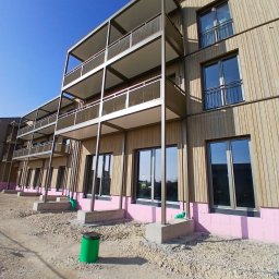 Sebastian Dobrowolski - Nowoczesny budynek z drewnianą elewacją i balkonami w trakcie budowy. Widoczne ocieplenie fundamentów i teren przygotowany pod zagospodarowanie.