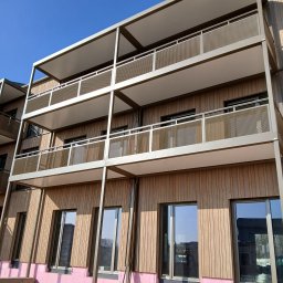 Sebastian Dobrowolski - Nowoczesny budynek z drewnianą elewacją, balkonami z metalową siatką i różową izolacją fundamentów, sfotografowany w słoneczny dzień.