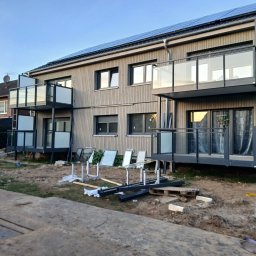 Sebastian Dobrowolski - Nowoczesny budynek z drewnianą elewacją i balkonami z szklanymi balustradami. Widoczne panele słoneczne na dachu i elementy konstrukcyjne na trawniku.