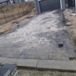 Sebastian Dobrowolski - Częściowo ułożona, szara kostka brukowa na podjeździe. Widoczne krawężniki, piasek i narzędzia. Etap prac brukarskich przed domem z bramą garażową.