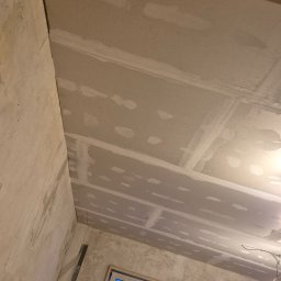 Kamil Perczak - Poddasze w trakcie ocieplania: widoczne płyty gipsowo-kartonowe na suficie, szpachlowane łączenia, surowe ściany i okna z niebieskim światłem dziennym. Rozstawiona drabina w rogu pokoju.